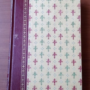 May include: A burgundy hardcover book with a gold-trimmed spine. The cover features a repeating pattern of green and red fleurs-de-lis on a cream background.