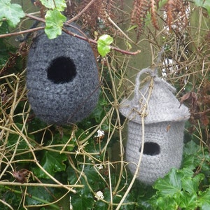 Könnte beinhalten: Zwei graue, gestrickte Vogelhäuschen hängen an einem mit Ranken bewachsenen Zaun. Die Vogelhäuschen haben runde Öffnungen, durch die die Vögel hineinfliegen können.