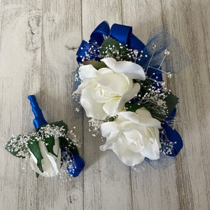 May include: A white rose corsage and boutonniere set with blue ribbon and accents. The corsage features three white roses, baby's breath, and blue netting. The boutonniere has a single white rose and blue ribbon.