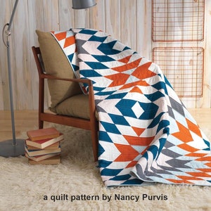 May include: A quilt pattern called "Four Corners" by Nancy Purvis, featuring a geometric design with orange, blue, white, and gray triangles. The quilt is draped over a wooden armchair in a room with a wooden wall and a lamp.