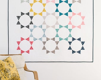 May include: A white quilt with a geometric pattern of circles and triangles in various colors, including teal, pink, gray, and yellow. The quilt is hanging on a white wall above a wooden chair with a patterned pillow.