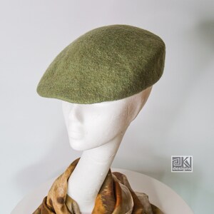 Puede incluir: Un gorro de fieltro verde sobre una cabeza de maniquí blanca. El maniquí lleva una bufanda con estampado marrón y beige.