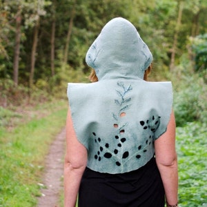 May include: A light teal hooded cape with cut-out leaf and dot designs. The cape is worn over a black dress, with a forest path in the background. The cape has a unique, artistic design.