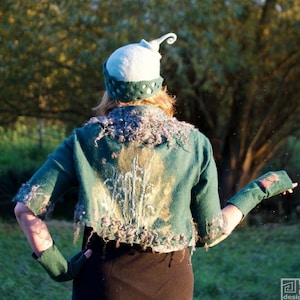 May include: A woman wearing a teal green felted jacket with a floral design, a light blue felted hat with a pointed top, and black gloves. The jacket has a frayed edge and the hat has a decorative design.