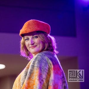 May include: A woman wearing an orange beret and a colourful shawl. The shawl features a blend of orange, grey, and yellow hues. The beret is a solid, vibrant orange colour. The background is a blurred purple.