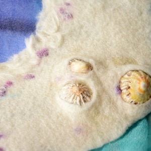 Op de afbeelding: Een witte viltstof met drie schelpen eraan bevestigd. De stof heeft een blauw en paars aquarelontwerp.