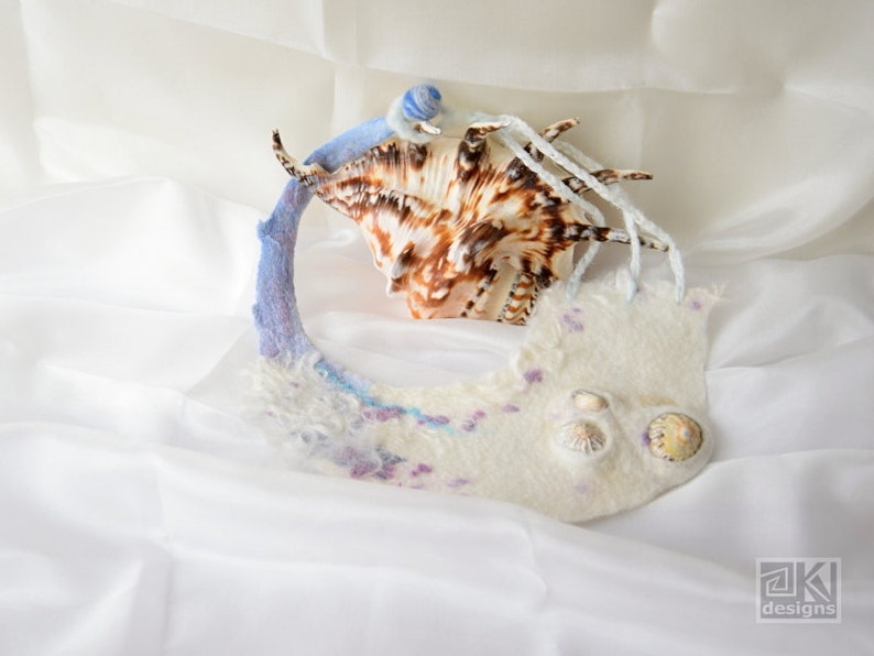 May include: A white felted fabric necklace with a seashell design. The necklace is decorated with small seashells and has a blue and purple gradient.