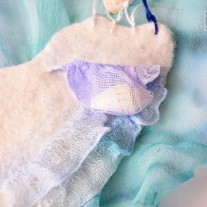 May include: A white felt heart with a blue and purple fabric ruffle and a seashell. The heart is on a blue and green fabric background.