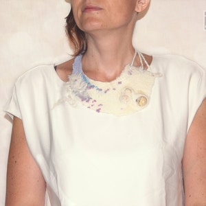 May include: A white, off-white, and light blue felted fabric necklace with a textured, organic shape. The necklace has a light blue, white, and purple color scheme.