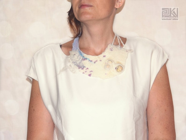May include: A white, off-white, and light blue felted fabric necklace with a textured, organic shape. The necklace has a light blue, white, and purple color scheme.