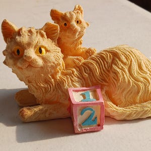 May include: A textured orange cat figurine featuring a smaller cat resting on its back. The larger cat has bright yellow eyes and is positioned next to a small pink cube block with the blue numbers 1 and 2 printed on its front and top faces.