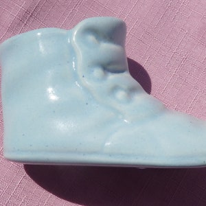 May include: A pale blue ceramic baby boot figurine. The boot features a high top with button detailing and a rounded toe. The figurine is a solid colour and sits on a pink fabric surface.