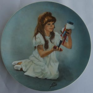 May include: Decorative plate featuring a young girl in a white dress holding a nutcracker toy. The plate has a light blue border and a painted portrait of the girl with long brown hair. The nutcracker is red, white, and blue.