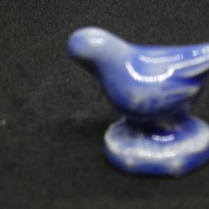 May include: A small, ceramic bird figurine in shades of blue and white. The bird is perched on a round base, with its wings slightly outstretched. The figurine is likely a decorative item.