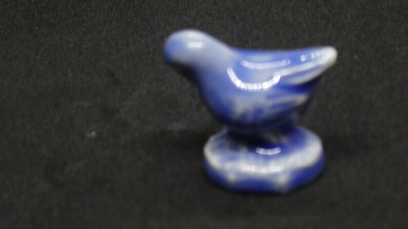 May include: A small, ceramic bird figurine in shades of blue and white. The bird is perched on a round base, with its wings slightly outstretched. The figurine is likely a decorative item.