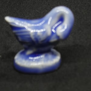 May include: A small, ceramic figurine of a swan in shades of blue and white. The swan has a curved neck and is perched on a round base. The figurine is likely a decorative item.