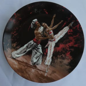 May include: Decorative plate featuring a ballet scene. The plate depicts a male and female dancer in white and red attire, set against a dark background with red accents. The dancers are in mid-motion, with the female dancer in a ballet pose.