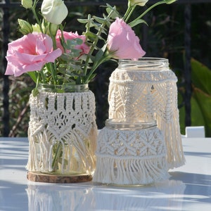 May include: Three glass jars with macrame covers, two are smaller and one is taller. The jars are filled with pink and white flowers.
