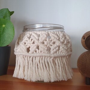 May include: A clear glass jar with a white crocheted cover and fringe. The jar is sitting on a wooden surface next to a small wooden figurine.