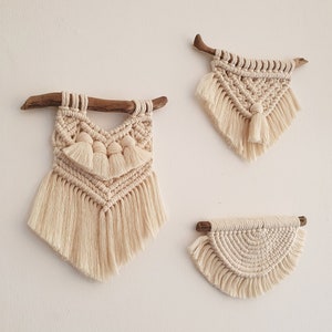 May include: Three macrame wall hangings with fringe, each with a different design. The hangings are made with natural coloured yarn and are attached to wooden branches.