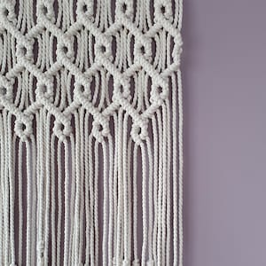 Puede incluir: Un tapiz de macramé blanco con un patrón geométrico y flecos. El tapiz está contra una pared de color púrpura claro.