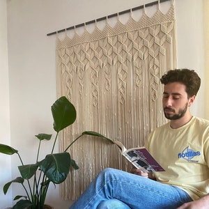 May include: A person sitting in a chair reading a book. They are wearing blue jeans and a yellow t-shirt with the text "flodllas" on it. The person is sitting in front of a macrame wall hanging.