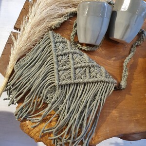 May include: A green macrame bag with fringe hanging from the bottom. The bag is on a wooden surface with two grey ceramic mugs.