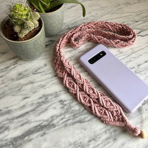 Puede incluir: Una correa de teléfono de crochet rosa con una funda de teléfono de color púrpura claro sobre una superficie de mármol blanco. La correa tiene un patrón de diamante y una cuenta de madera en el extremo.