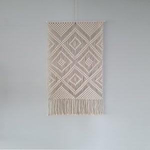 Könnte beinhalten: Ein weißer Makramee-Wandbehang mit einem Diamantenmuster und Fransen.