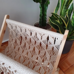 Può includere: Un arazzo da parete in macramè di legno di colore beige naturale. Il macramè è realizzato con un motivo a nodi e ha una frangia nella parte inferiore. L'arazzo è appeso a una parete bianca.