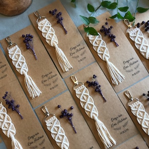 May include: A set of eight white macrame keychains with tassels, each attached to a brown card with the text "Derya'nin Kina Hatiras" and a date. The cards are decorated with dried purple flowers.