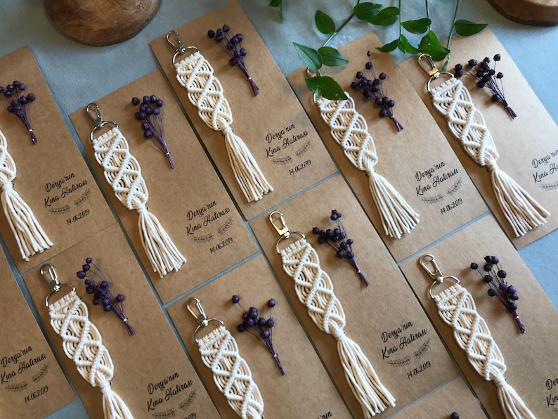 May include: A set of eight white macrame keychains with tassels, each attached to a brown card with the text "Derya'nin Kina Hatiras" and a date. The cards are decorated with dried purple flowers.