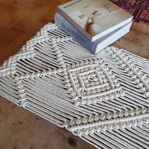 May include: A white macrame table runner with a geometric pattern. The runner is on a wooden table with books stacked on top of it. The books are titled "Interior Architecture", "Swedish Architecture", and "Eliasson".