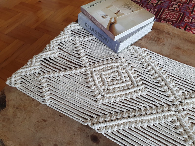 May include: A white macrame table runner with a geometric pattern. The runner is on a wooden table with books stacked on top of it. The books are titled "Interior Architecture", "Swedish Architecture", and "Eliasson".