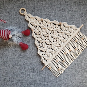 Peut inclure: Une suspension murale en macramé blanche en forme de triangle avec des perles en bois. La suspension a une frange en bas et est fixée à une baguette en bois.