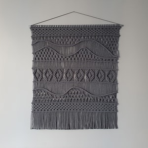 May include: Gray macrame wall hanging with a geometric pattern. The hanging has a fringe at the bottom.