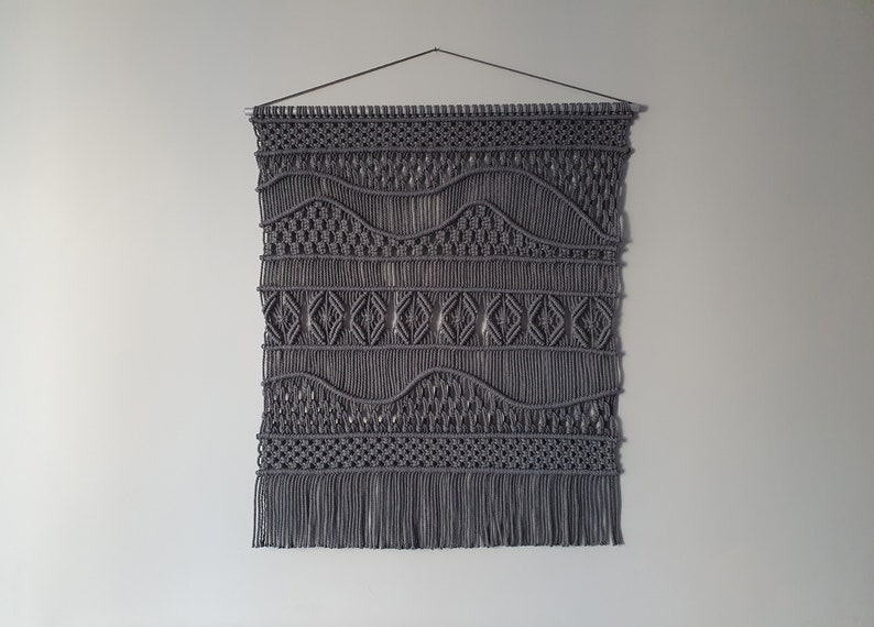 May include: Gray macrame wall hanging with a geometric pattern. The hanging has a fringe at the bottom.