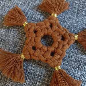 May include: A brown crocheted star with gold trim and tassels. The star is made of yarn and has a hole in the center.