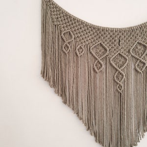 May include: A macrame wall hanging with a geometric pattern and fringe. The hanging is made of natural coloured cord and is attached to a wooden ring.