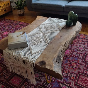 May include: A wooden coffee table with a live edge design and a white macrame table runner. The table is sitting on a red and purple patterned rug. The book "Interior Architecture" is on the table.