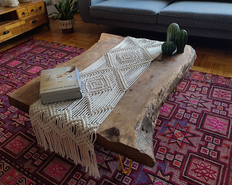 May include: A wooden coffee table with a live edge design and a white macrame table runner. The table is sitting on a red and purple patterned rug. The book "Interior Architecture" is on the table.