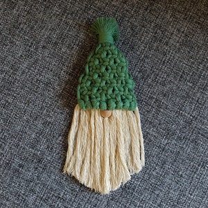 May include: A green crocheted gnome with a long, white beard. The gnome is wearing a green hat with a white pom-pom.