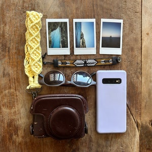 Puede incluir: Una disposición plana de artículos esenciales para viajar que incluye una funda para cámara, unas gafas de sol, una funda para teléfono, una pulsera de cuentas, una correa de cámara de macramé amarilla y tres fotos instantáneas.
