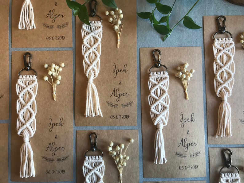 May include: A set of six white macrame keychains with a tassel on the bottom. Each keychain is attached to a brown card with the text "Jpek & Alper" and the date "06.04.2019".