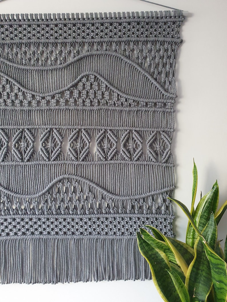 May include: Gray macrame wall hanging with a geometric pattern and fringe. The hanging is made with thick cotton cord and has a woven design.