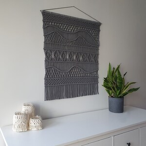 May include: Gray macrame wall hanging with a geometric pattern. The wall hanging is made of thick cord and has a fringe at the bottom. The wall hanging is hanging on a white wall above a white dresser with three drawers.