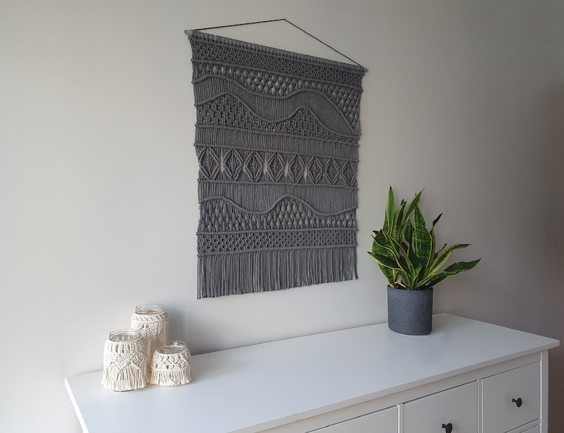May include: Gray macrame wall hanging with a geometric pattern. The wall hanging is made of thick cord and has a fringe at the bottom. The wall hanging is hanging on a white wall above a white dresser with three drawers.