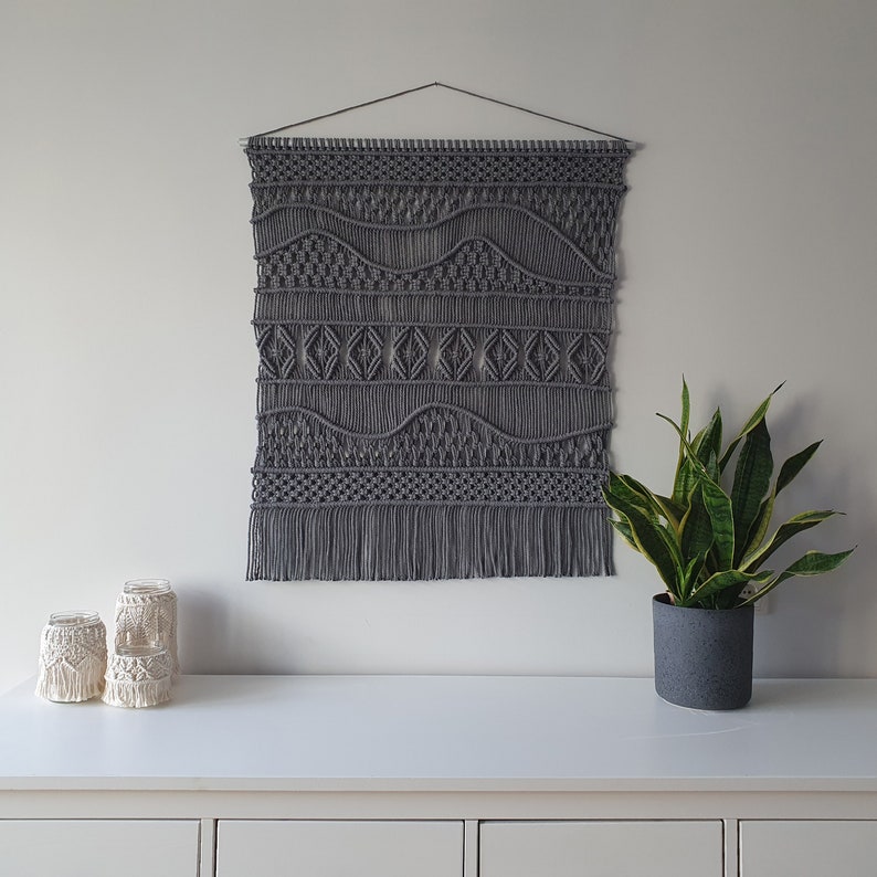 May include: Gray macrame wall hanging with a geometric pattern and fringe. The hanging is displayed on a white wall with a potted plant and candles on a white shelf below.