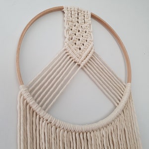 May include: A white macrame wall hanging with a wooden hoop and a fringe. The macrame is in a geometric pattern.
