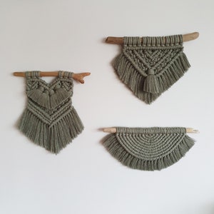 May include: Three macrame wall hangings with wooden dowels. The hangings are in a sage green color and have fringe details. The top hanging is a triangle shape, the middle hanging is a heart shape, and the bottom hanging is a half-circle shape.
